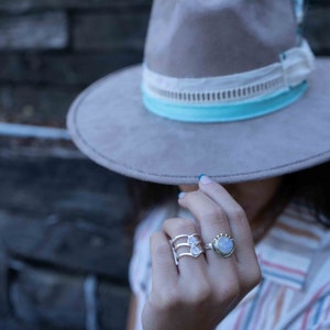 May include: A woman wearing a gray felt hat with a white and blue trim, and a striped shirt. She is wearing two silver rings with moonstone settings on her ring finger.