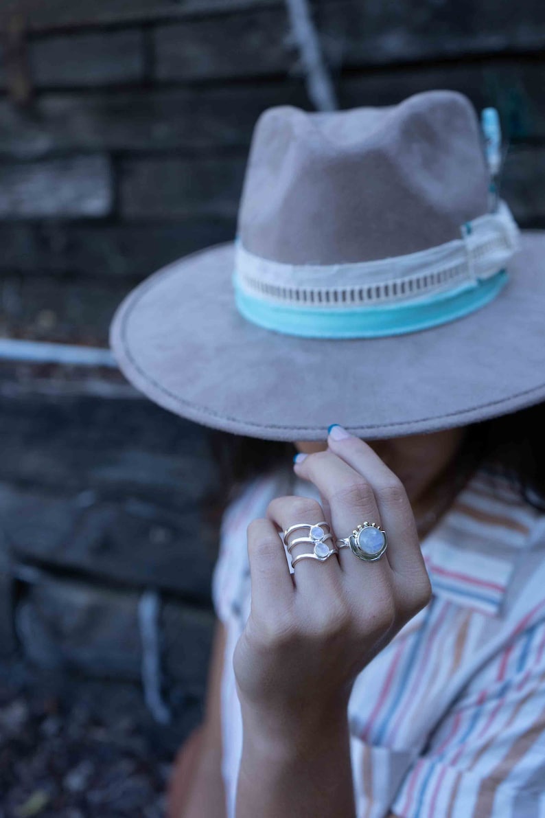May include: A woman wearing a gray felt hat with a white and blue trim, and a striped shirt. She is wearing two silver rings with moonstone settings on her ring finger.