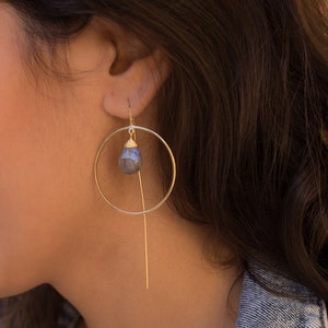 May include: Gold hoop earrings with a dangling teardrop-shaped gemstone. The gemstone is a dark grey colour with a slight iridescent sheen.