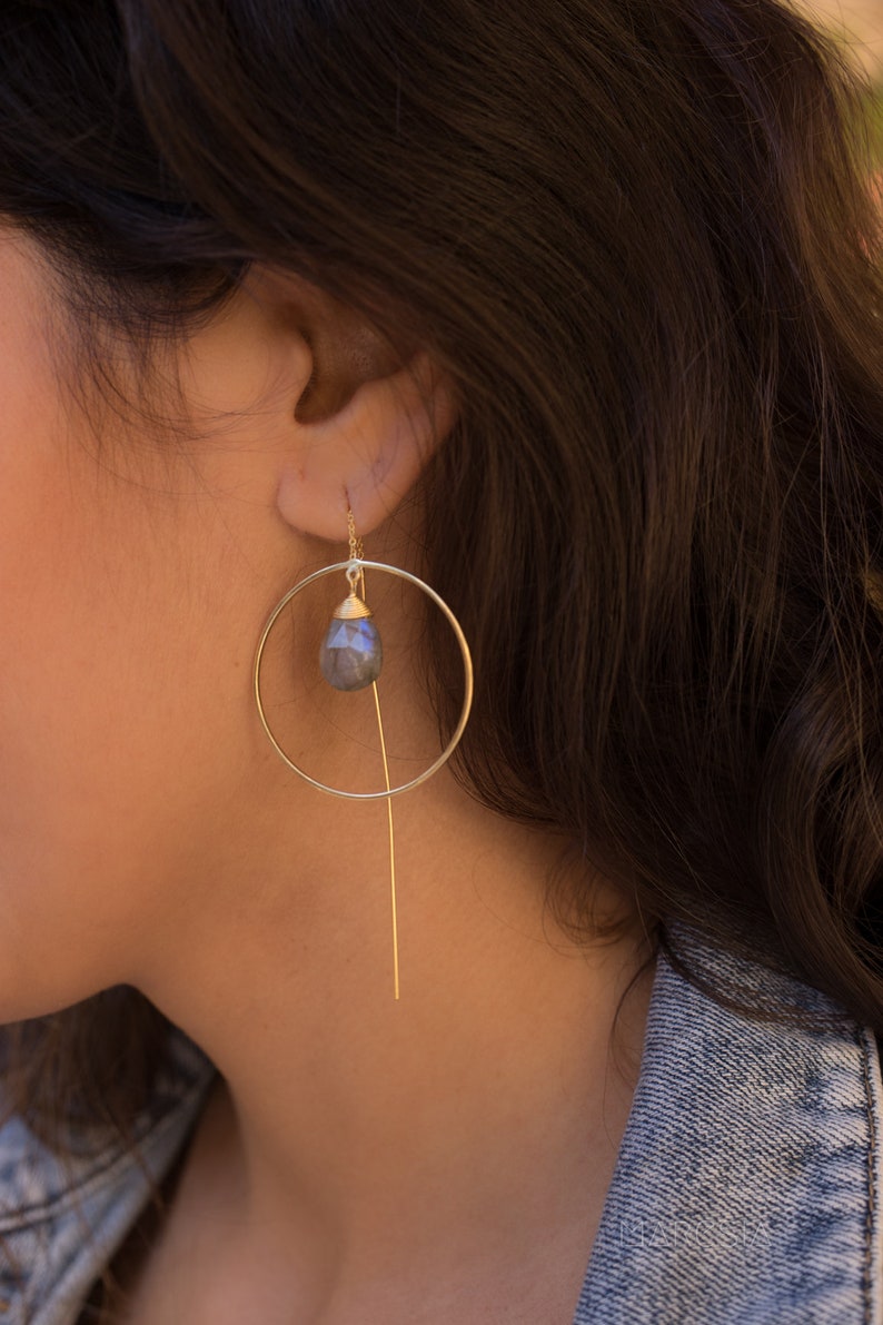 May include: Gold hoop earrings with a dangling teardrop-shaped gemstone. The gemstone is a dark grey colour with a slight iridescent sheen.