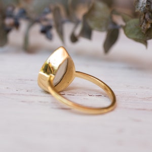 May include: A gold ring with a white oval-shaped gemstone. The ring is set on a white wooden surface.
