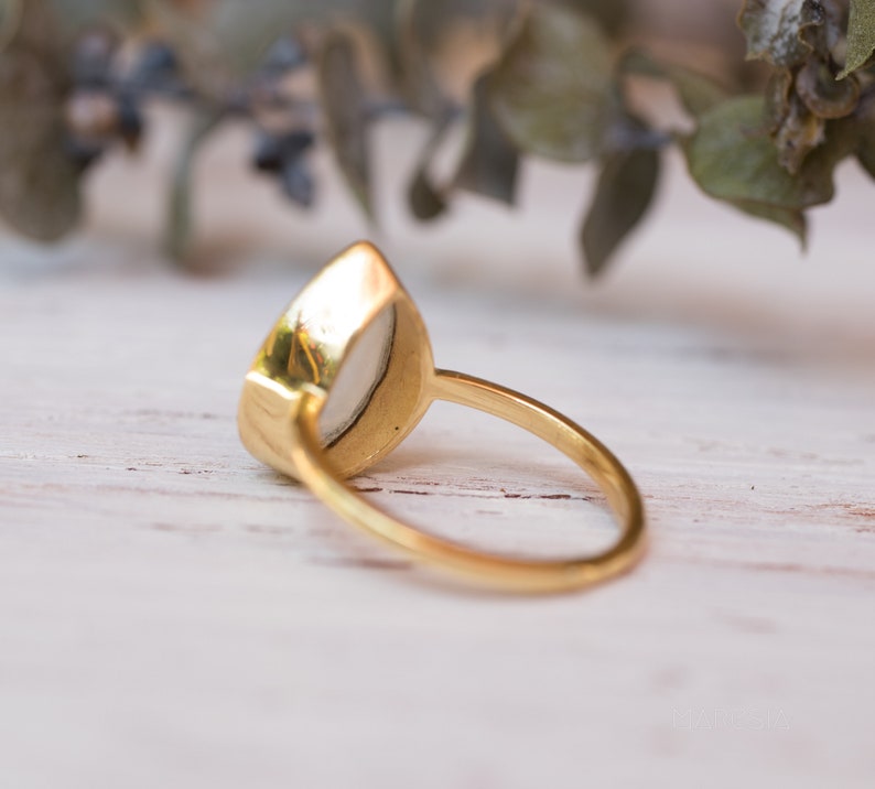 May include: A gold ring with a white oval-shaped gemstone. The ring is set on a white wooden surface.