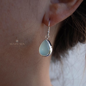 Peut inclure: Boucle d'oreille unique en forme de goutte d'eau avec une pierre bleu pâle et un cadre en métal argenté. La boucle d'oreille est suspendue à un crochet argenté. Le mot "MARESIA" est visible dans l'image.