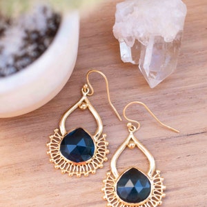 May include: A pair of gold dangle earrings with a dark blue gemstone in the center of each earring. The earrings have a delicate, intricate design.