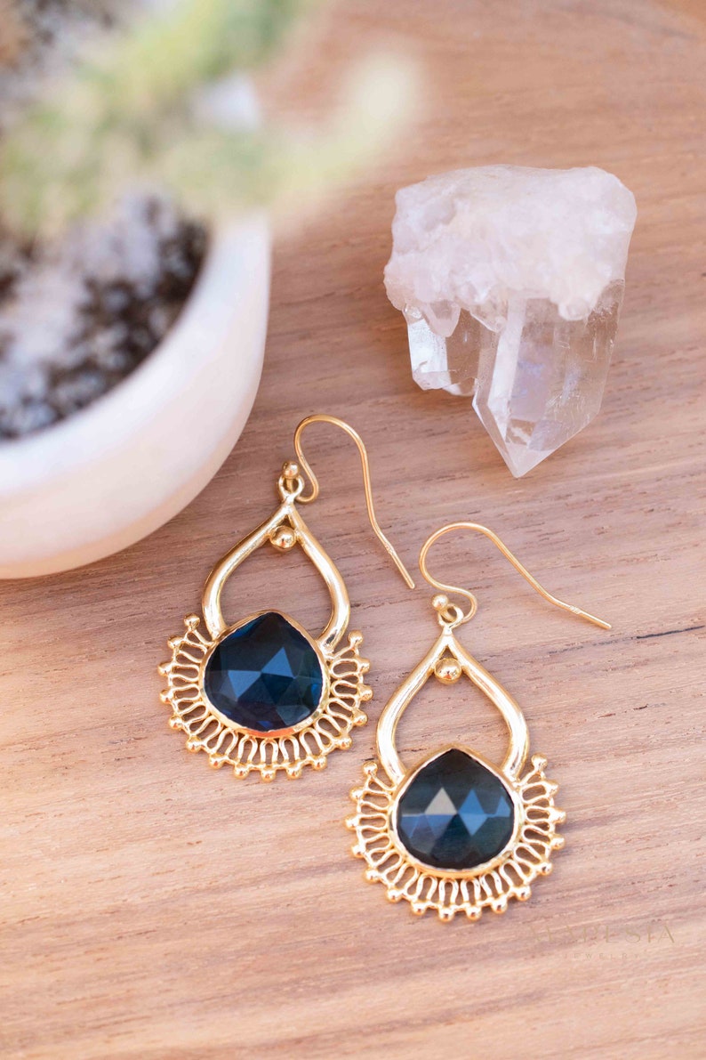 May include: A pair of gold dangle earrings with a dark blue gemstone in the center of each earring. The earrings have a delicate, intricate design.