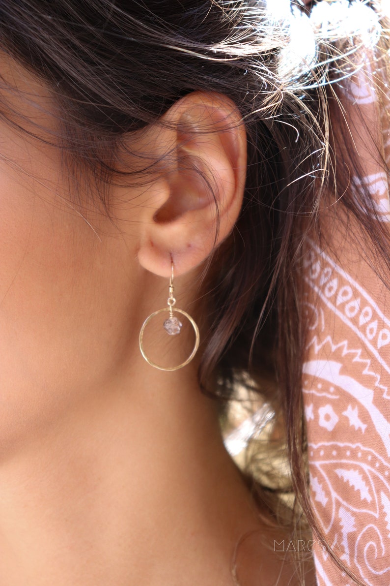 May include: Gold hoop earrings with a small clear crystal hanging from the bottom. The earrings are worn by a person with long brown hair.