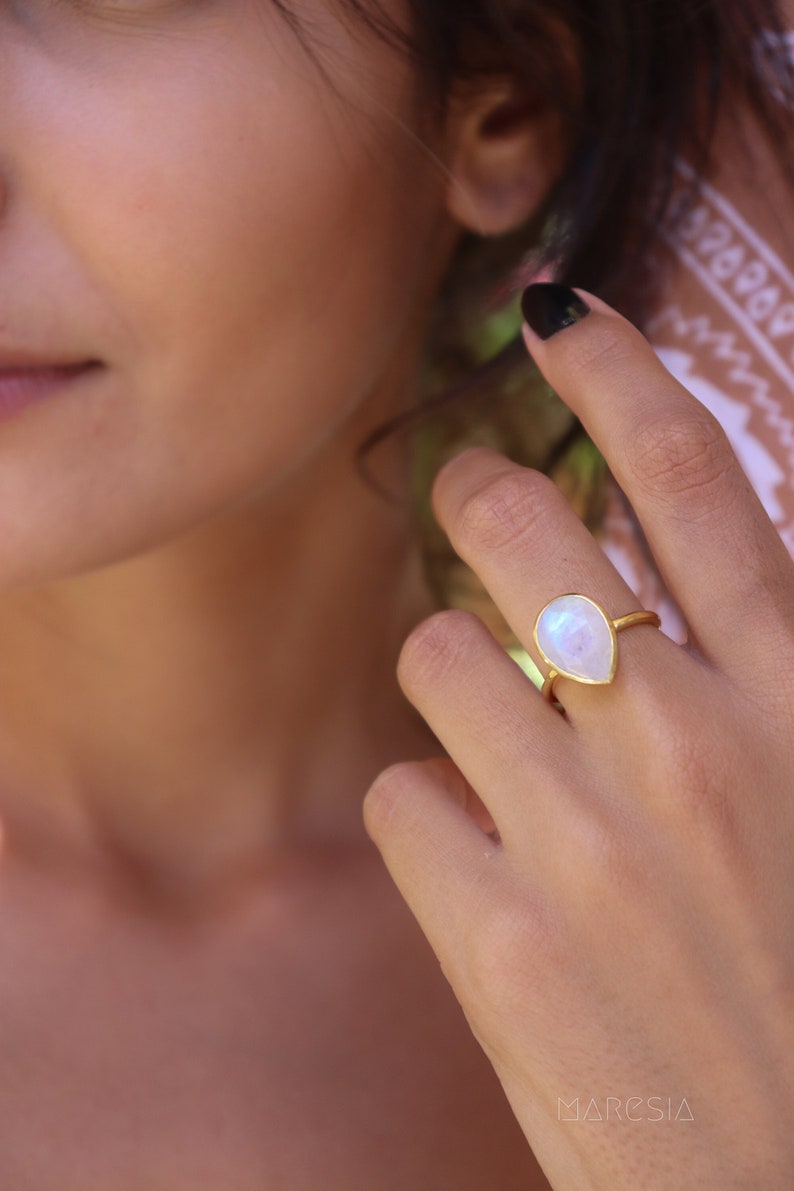 May include: A gold ring with a teardrop-shaped moonstone. The ring is on a person's finger.