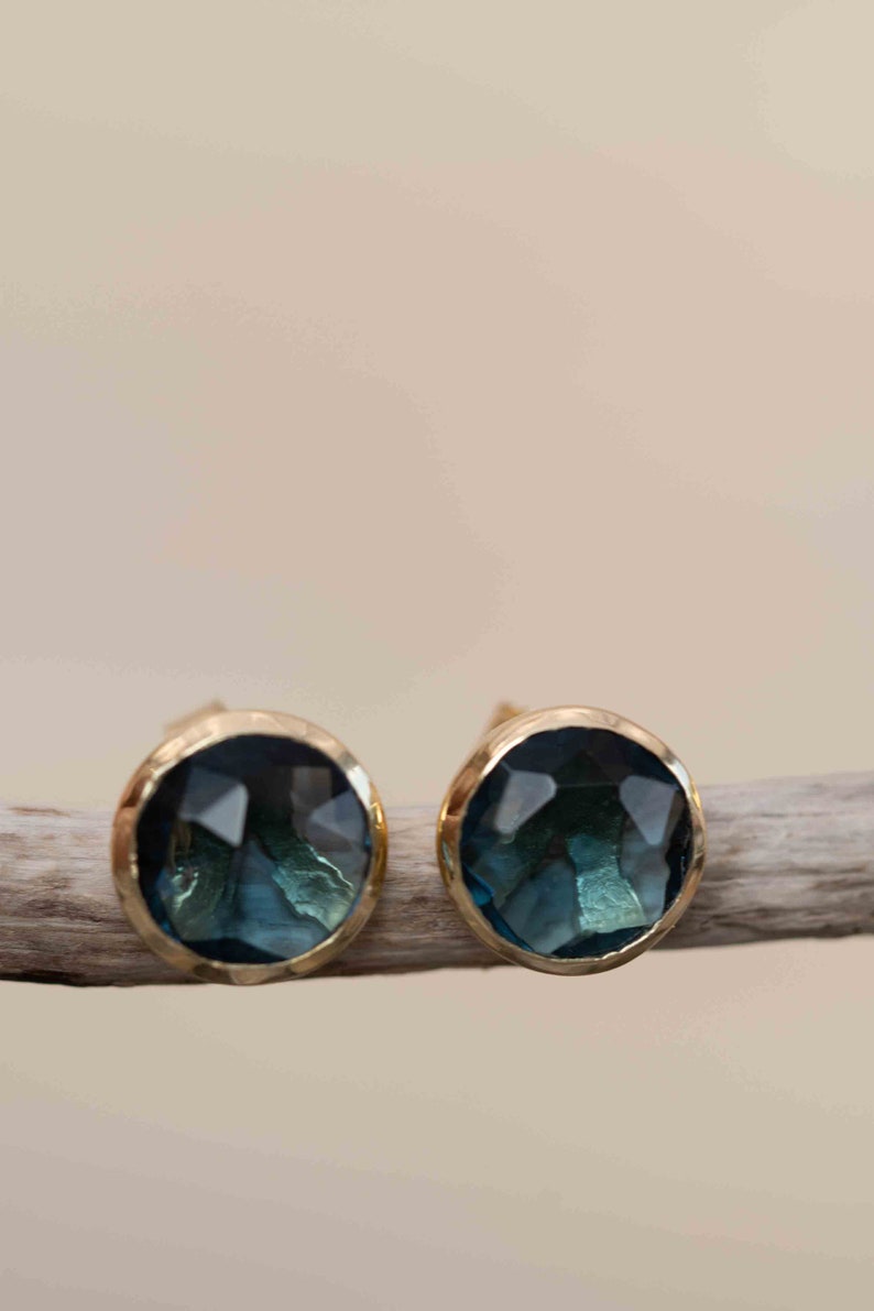 May include: A pair of gold stud earrings with a dark blue gemstone in each. The gemstones are faceted and have a slightly textured surface.