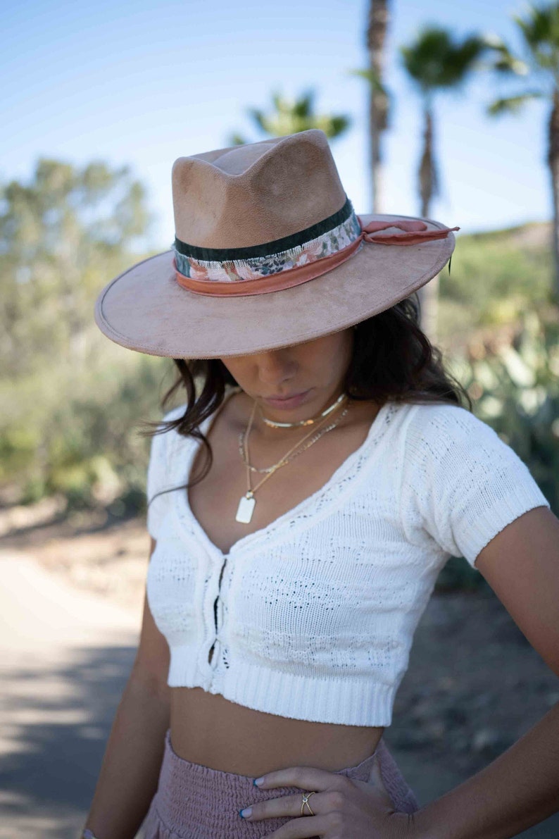 May include: A light brown fedora hat with a decorative band featuring floral and green accents. The hat has a wide brim and is worn with a white knit top and gold necklaces. The overall aesthetic is bohemian and stylish.
