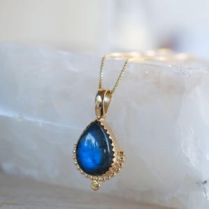 May include: A gold pendant necklace featuring a teardrop-shaped labradorite stone. The blue-grey stone is set in a gold bezel with a beaded edge. The necklace has a delicate gold chain.