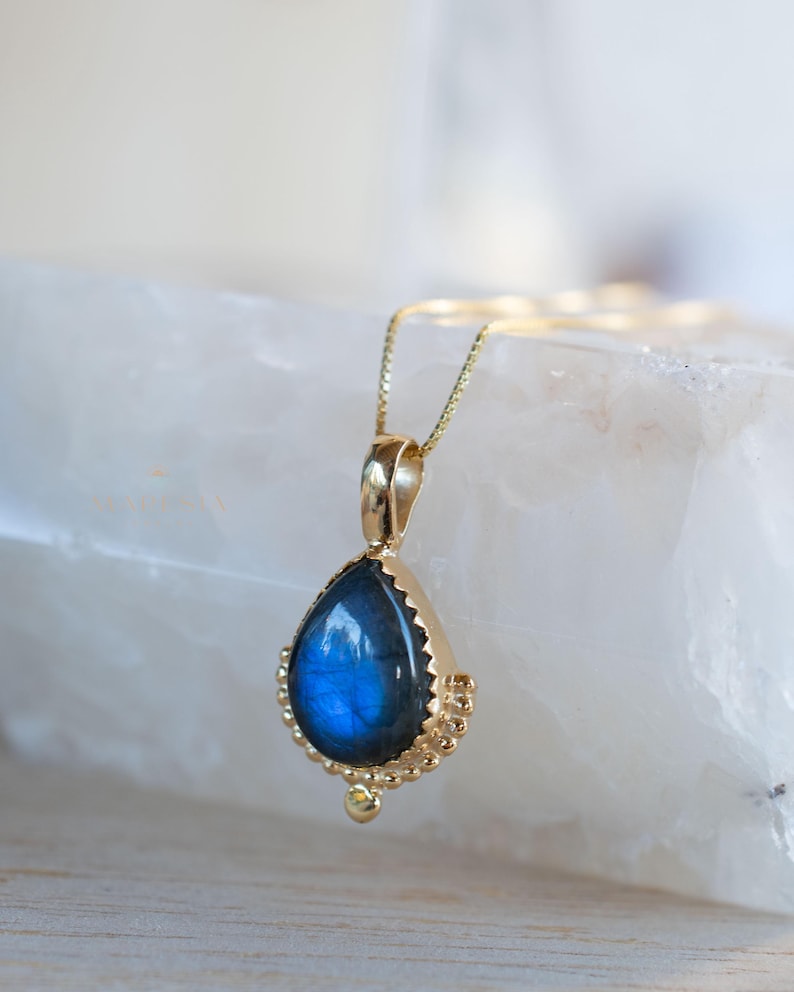 May include: A gold pendant necklace featuring a teardrop-shaped labradorite stone. The blue-grey stone is set in a gold bezel with a beaded edge. The necklace has a delicate gold chain.