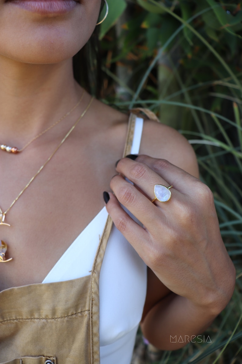May include: A gold ring with a teardrop-shaped moonstone. The ring is worn on the index finger of a person's hand.
