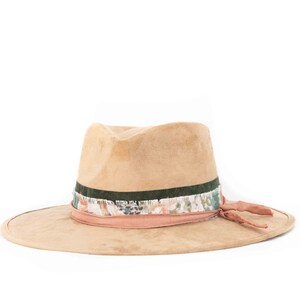 May include: A tan fedora hat with a wide brim. The hat features a dark green band, a patterned band with floral accents, and a pink band with a matching tie. The hat is made of a soft, suede-like material.