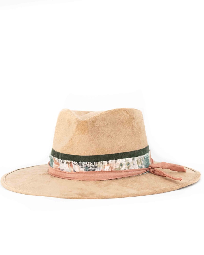 May include: A tan fedora hat with a wide brim. The hat features a dark green band, a patterned band with floral accents, and a pink band with a matching tie. The hat is made of a soft, suede-like material.