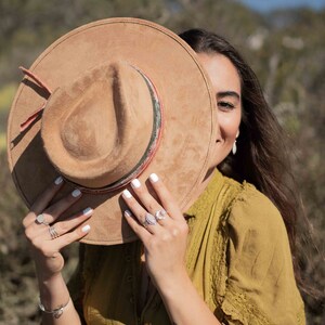May include: A tan suede hat with a decorative band and a red strap. The hat is held up by a person wearing a mustard-colored dress and silver rings. The background is a blurred outdoor scene.