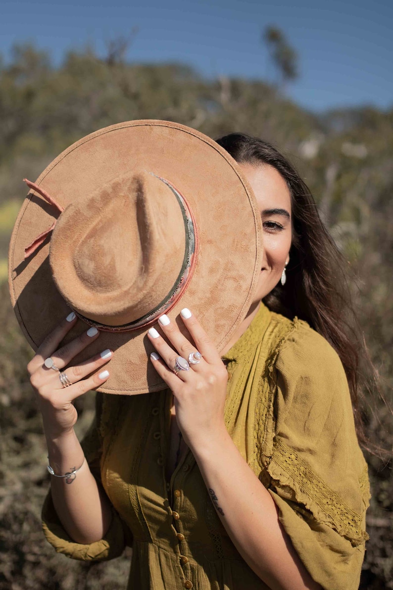 May include: A tan suede hat with a decorative band and a red strap. The hat is held up by a person wearing a mustard-colored dress and silver rings. The background is a blurred outdoor scene.