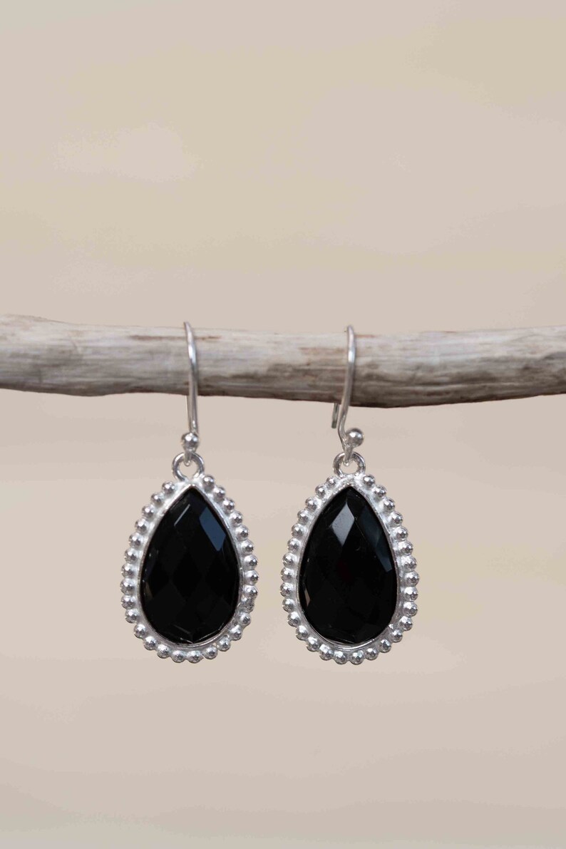 May include: A pair of silver earrings with black teardrop-shaped gemstones. The earrings have a beaded border around the gemstones.