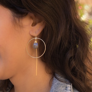 May include: Gold hoop earrings with a dangling, faceted, grey gemstone. The hoop is open and the gemstone hangs from a thin gold bar.