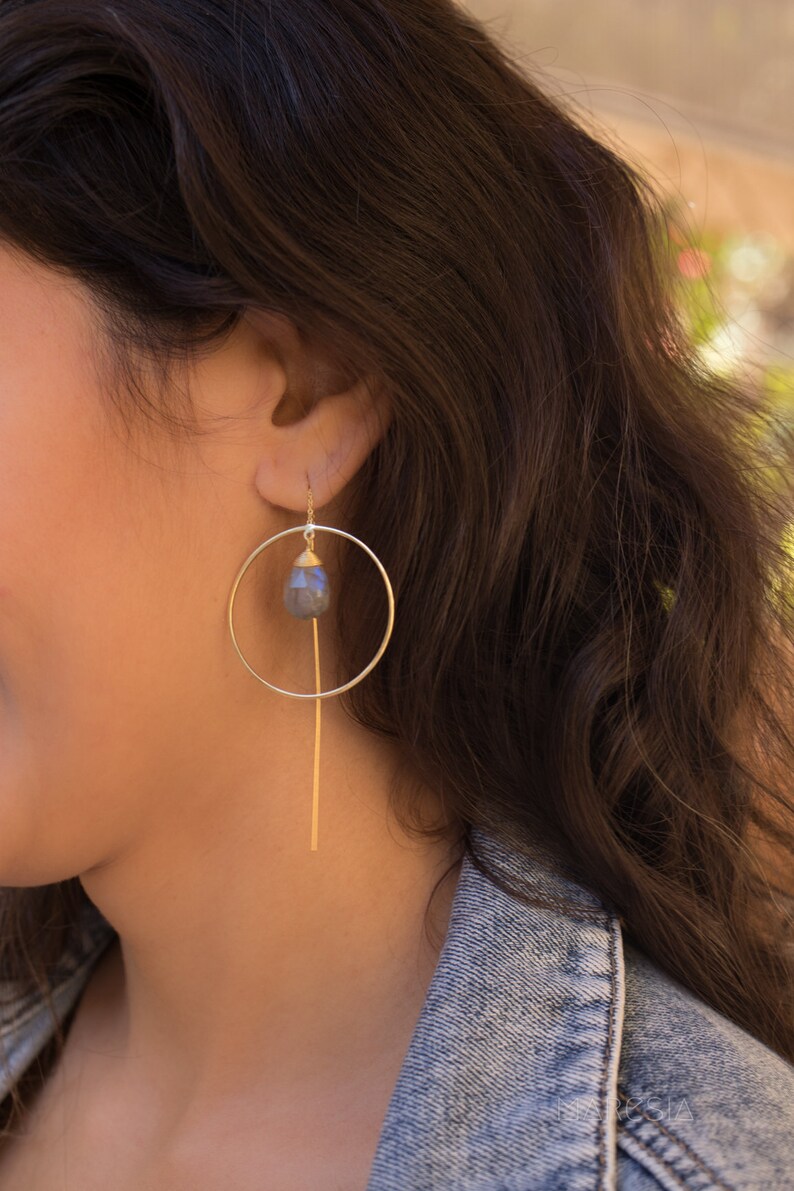 May include: Gold hoop earrings with a dangling, faceted, grey gemstone. The hoop is open and the gemstone hangs from a thin gold bar.