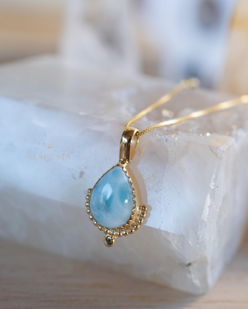 May include: A gold necklace with a teardrop-shaped pendant. The pendant features a light blue gemstone with a gold beaded border. The necklace is displayed on a white surface. The word "MARESIA" is visible in the background.