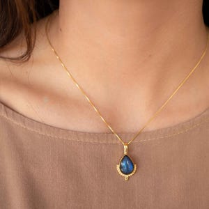 May include: A gold-colored necklace with a teardrop-shaped pendant. The pendant features a blue gemstone with a gold-colored decorative border. The necklace is worn against a neutral-colored top.
