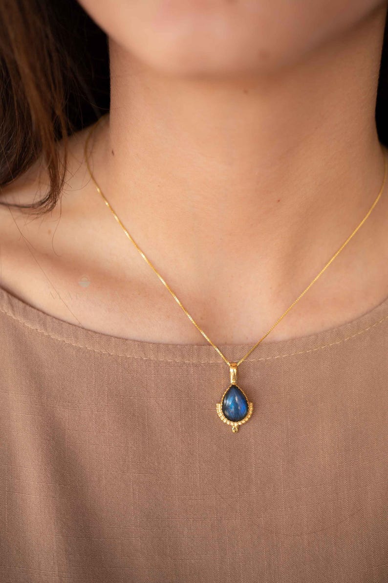 May include: A gold-colored necklace with a teardrop-shaped pendant. The pendant features a blue gemstone with a gold-colored decorative border. The necklace is worn against a neutral-colored top.