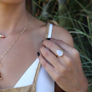 May include: A gold ring with a teardrop-shaped moonstone. The ring is worn on the index finger of a person's hand.