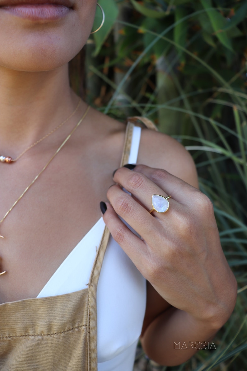 May include: A gold ring with a teardrop-shaped moonstone. The ring is worn on the index finger of a person's hand.