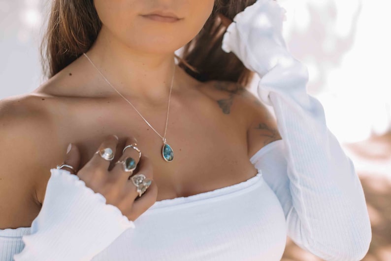 May include: A woman wearing a white off-the-shoulder top with a silver necklace and a teardrop-shaped blue gemstone pendant. She is wearing several silver rings on her fingers.