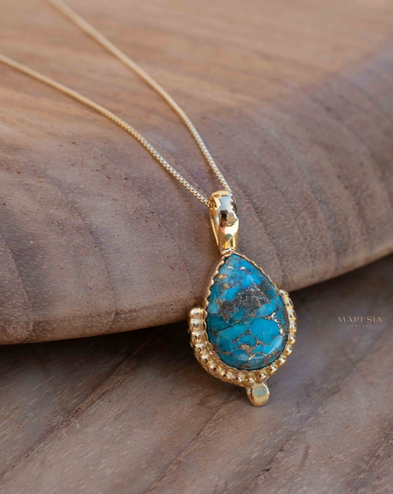 May include: A gold-colored necklace with a teardrop-shaped pendant. The pendant features a vibrant turquoise stone with gold flecks, set in a detailed gold bezel. The necklace is displayed on a wooden surface.