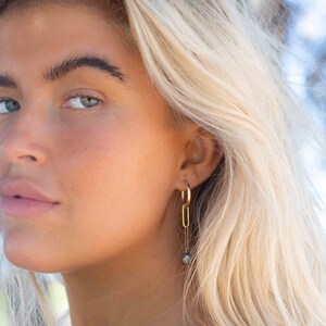 K&ouml;nnte beinhalten: Eine Frau mit blonden Haaren tr&auml;gt goldene Creolen mit einem baumelnden schwarzen Perlenanh&auml;nger.