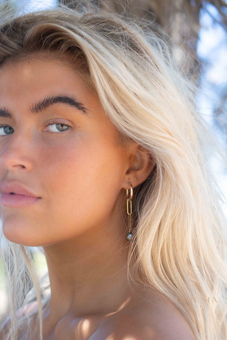 K&ouml;nnte beinhalten: Eine Frau mit blonden Haaren tr&auml;gt goldene Creolen mit einem baumelnden schwarzen Perlenanh&auml;nger.