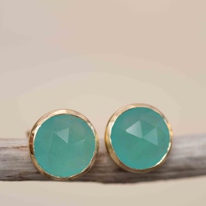 May include: A pair of gold earrings with teal gemstone studs. The earrings are on a light brown wooden surface.