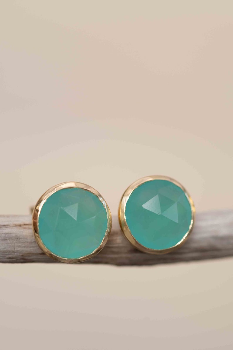 May include: A pair of gold earrings with teal gemstone studs. The earrings are on a light brown wooden surface.