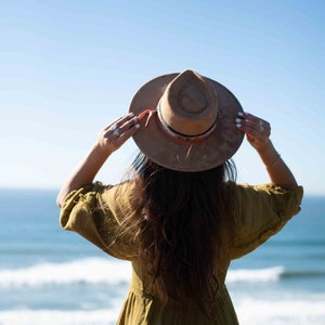 May include: A tan fedora-style hat with a decorative band is held up against a bright blue sky. The hat is worn with a long-sleeved olive green dress. The ocean is visible in the background.