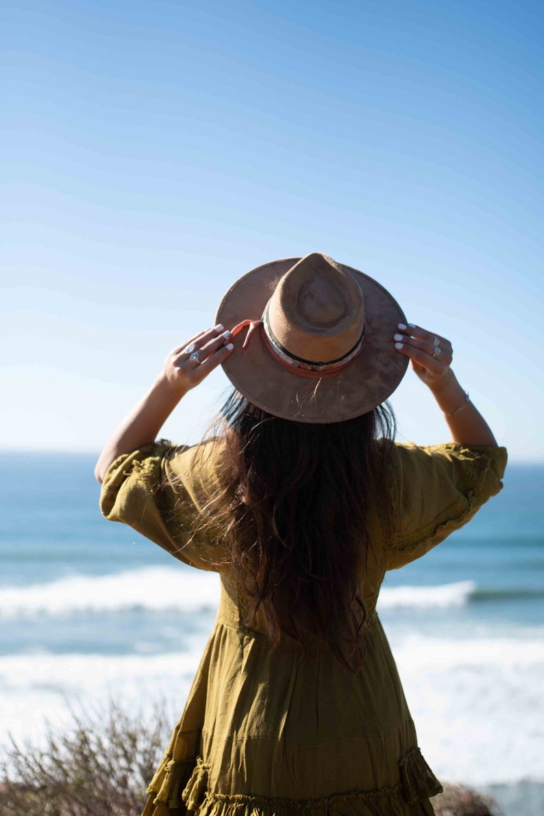 May include: A tan fedora-style hat with a decorative band is held up against a bright blue sky. The hat is worn with a long-sleeved olive green dress. The ocean is visible in the background.