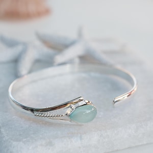 May include: A silver bangle bracelet with a light blue gemstone. The bracelet is open and has a delicate design.