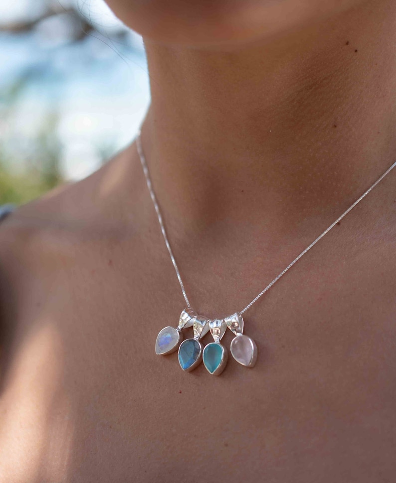 May include: A silver necklace with five teardrop-shaped pendants. The pendants are made of different gemstones, including moonstone, labradorite, aquamarine, and rose quartz.