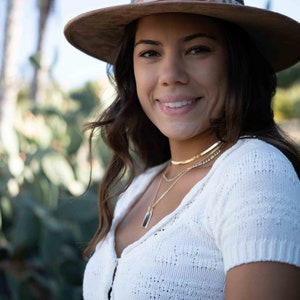 May include: A brown fedora hat with a decorative band featuring a floral pattern. The hat is worn with a white knit top and gold necklaces. The image is taken outdoors with a blurred background of green foliage.