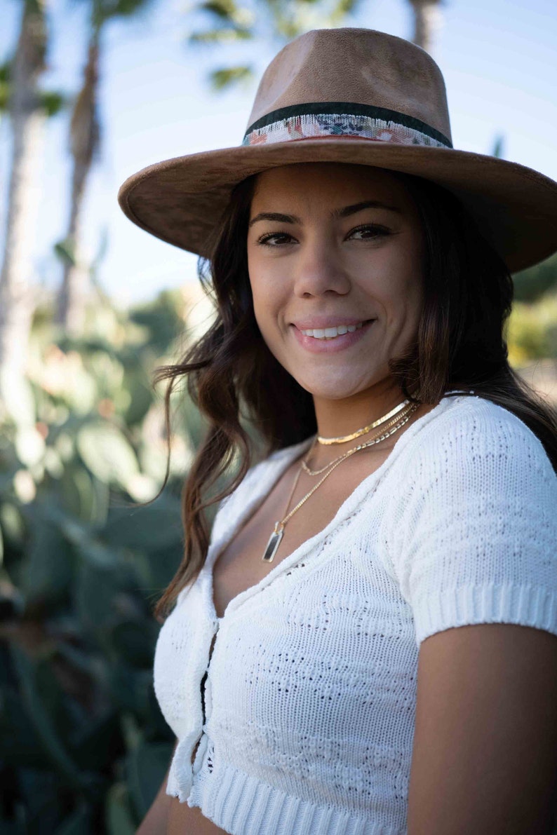 May include: A brown fedora hat with a decorative band featuring a floral pattern. The hat is worn with a white knit top and gold necklaces. The image is taken outdoors with a blurred background of green foliage.