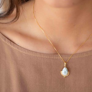 May include: A gold-colored necklace with a teardrop-shaped pendant. The pendant features a light blue gemstone with a decorative gold setting. The necklace is displayed against a neutral background.