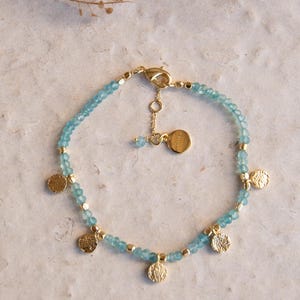May include: A delicate gold bracelet featuring small, light blue beads and several gold coin charms. The bracelet has a lobster clasp closure and an adjustable chain. The bracelet is displayed on a neutral surface.