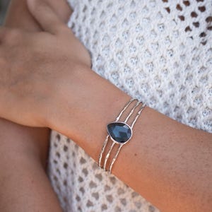 May include: A silver-toned cuff bracelet featuring three thin, textured bands that converge at a central teardrop-shaped dark gray stone. The metal bands have a hammered or irregular finish, and the stone is set in a smooth, polished silver-colored frame.