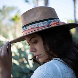 May include: A tan fedora hat with a wide brim. The hat features a decorative band with floral and peach-colored accents. The hat is worn by a person in a white sweater, with a blurred green background.