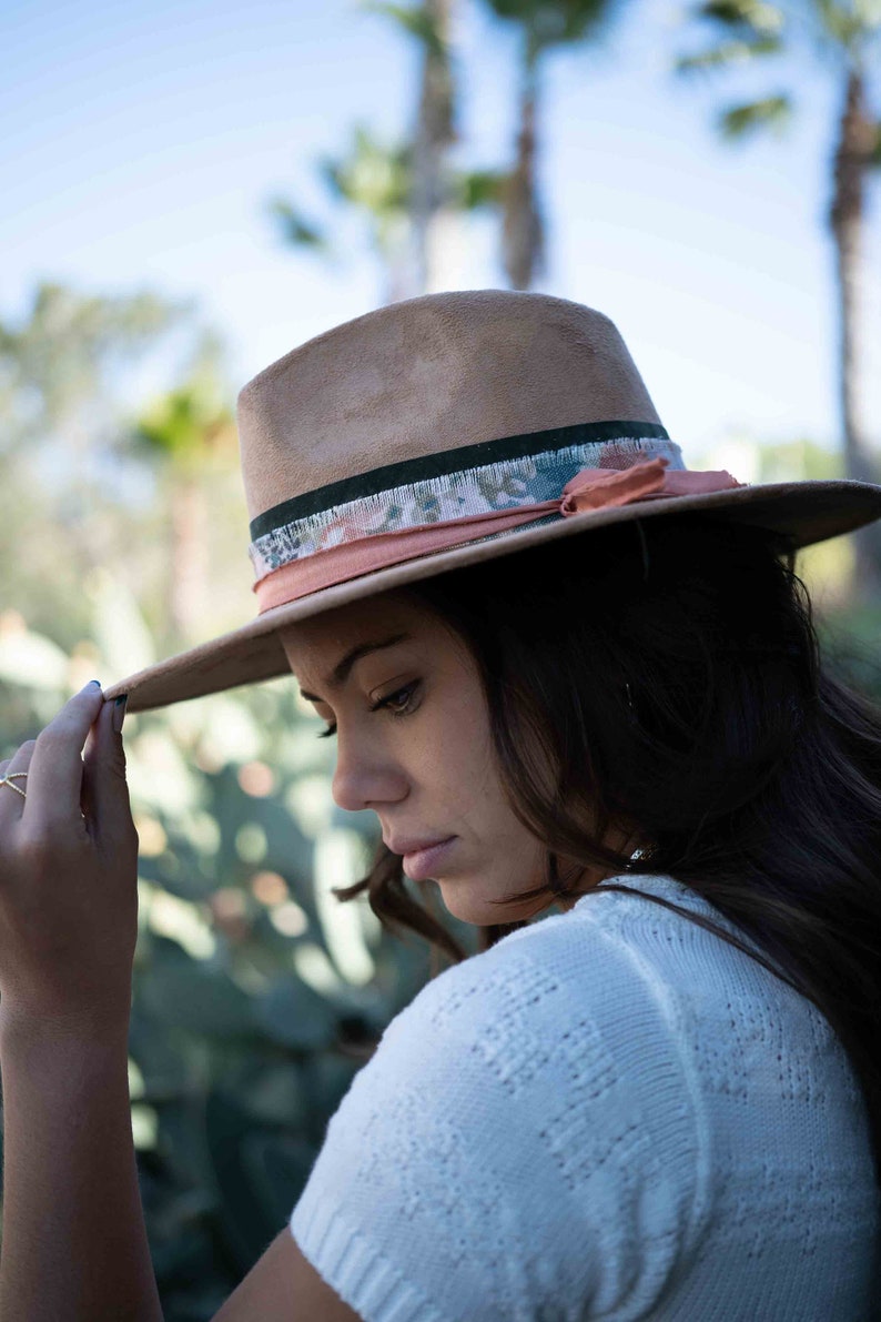 May include: A tan fedora hat with a wide brim. The hat features a decorative band with floral and peach-colored accents. The hat is worn by a person in a white sweater, with a blurred green background.