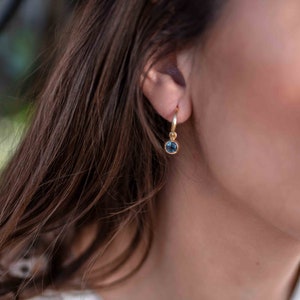 May include: A gold hoop earring with a dangling blue gemstone. The earring is worn in the left ear of a person with long brown hair.