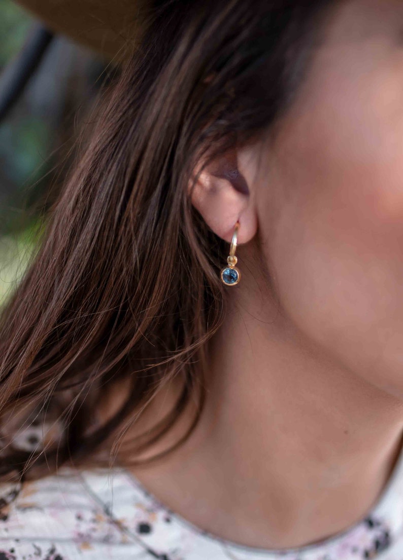 May include: A gold hoop earring with a dangling blue gemstone. The earring is worn in the left ear of a person with long brown hair.