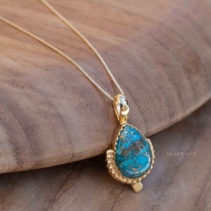 May include: A gold-colored necklace with a teardrop-shaped pendant. The pendant features a vibrant turquoise stone with brown veining, set in a textured gold frame. The necklace is displayed on a wooden surface.