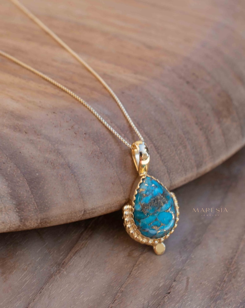 May include: A gold-colored necklace with a teardrop-shaped pendant. The pendant features a vibrant turquoise stone with brown veining, set in a textured gold frame. The necklace is displayed on a wooden surface.
