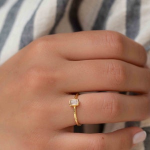 May include: A gold ring with a rectangular clear gemstone. The ring is on a finger.